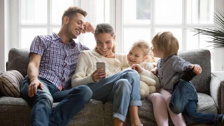 Mom, dad, and two kids smiling look at a phone on a couch, los gatos ca
